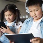 Children using smartphone and tablet on sofa, boy wearing headphones while girl plays on mobile device