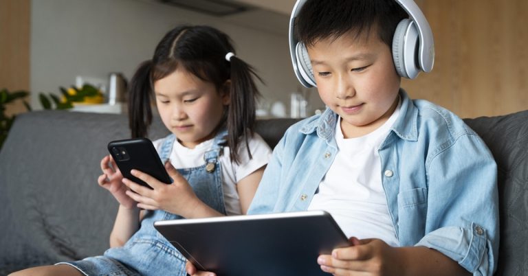 Children using smartphone and tablet on sofa, boy wearing headphones while girl plays on mobile device