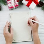 Hands writing in notebook on white table with Christmas gifts, pine branches and holiday decorations