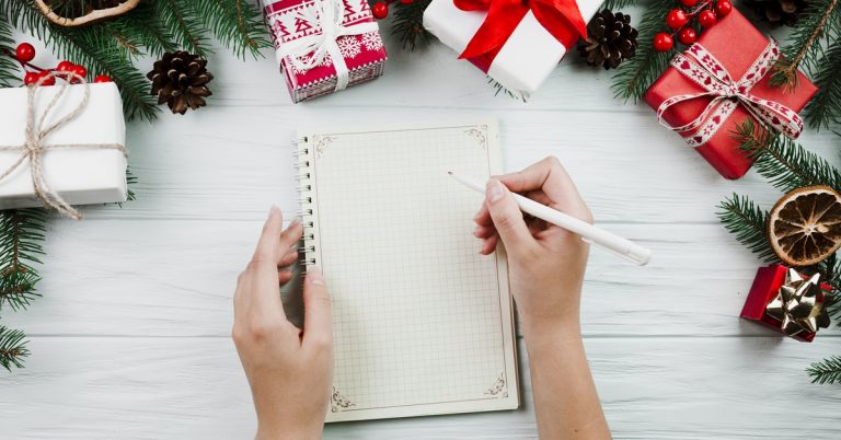 Hands writing in notebook on white table with Christmas gifts, pine branches and holiday decorations