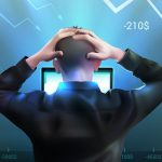 Stressed trader holding head while looking at computer as stock market charts fall and losses are shown on screen