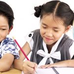 Two children studying together at a desk, smiling while writing in a notebook with a globe nearby