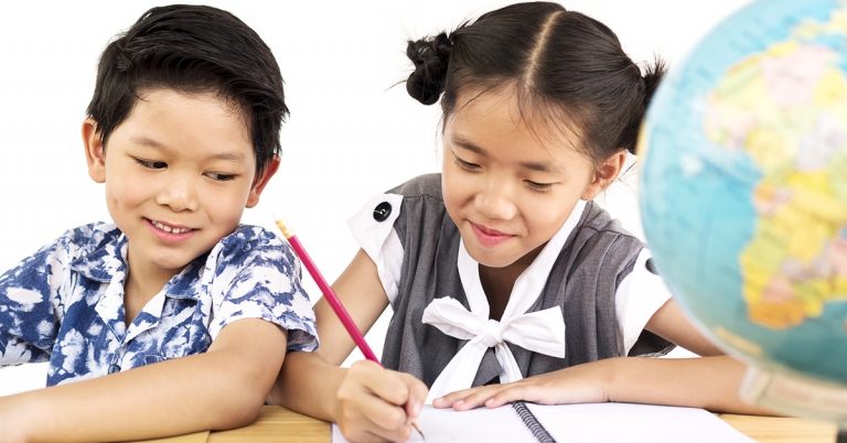 Two children studying together at a desk, smiling while writing in a notebook with a globe nearby