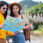 Two female tourists reading a world map outdoors, planning travel near a river and bridge