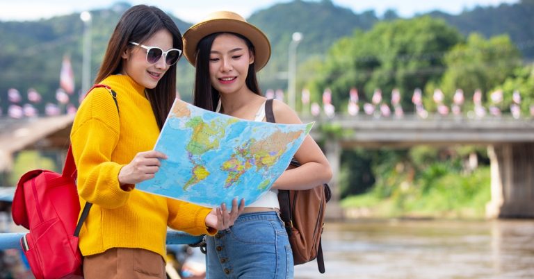 Two female tourists reading a world map outdoors, planning travel near a river and bridge