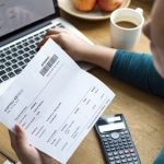 Person reviewing shipping invoice and expenses at desk with laptop, calculator, coffee and smartphone