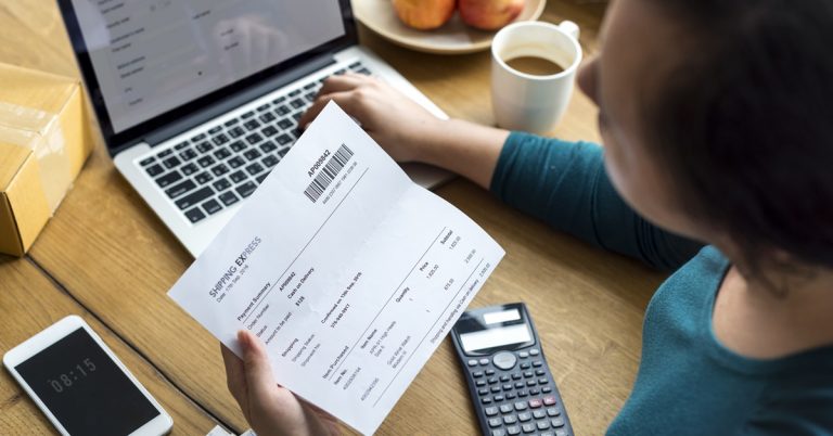 Person reviewing shipping invoice and expenses at desk with laptop, calculator, coffee and smartphone