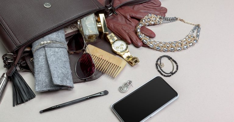 Contents of a women’s handbag including jewelry, watch, gloves, sunglasses, makeup brush, wallet and smartphone on table