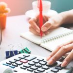 Person calculating expenses with calculator while writing in notebook, budgeting finances at a desk