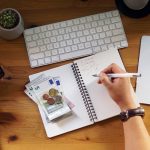 Person calculating personal finances at desk with calculator, notebook, cash, keyboard and coffee