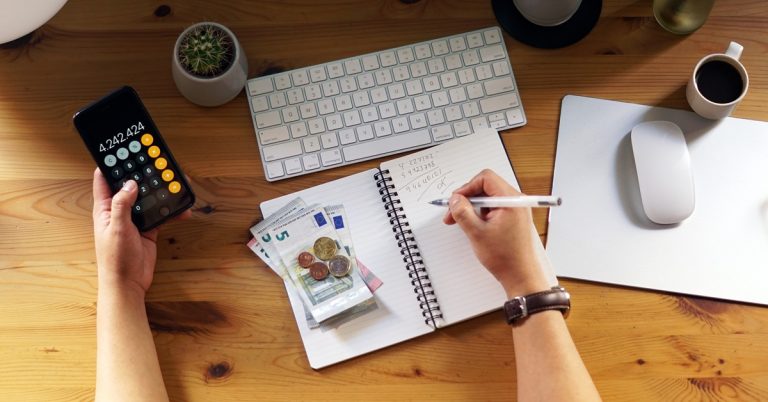 Person calculating personal finances at desk with calculator, notebook, cash, keyboard and coffee