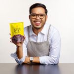 Smiling man holding a spicy beef jerky snack pack, presenting a Malaysian food product in a studio setting