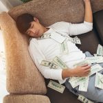 Man sleeping on sofa covered with dollar bills at home, symbolizing wealth, financial success or money windfall