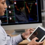 Trader analyzing stock charts on tablet with market data displayed on computer screen in office setting