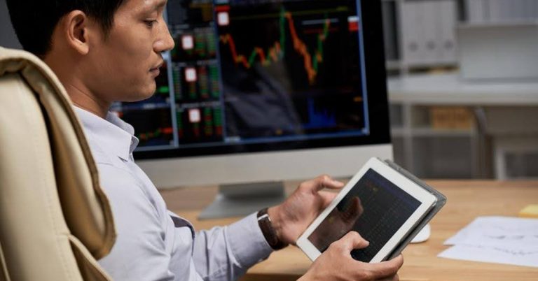 Trader analyzing stock charts on tablet with market data displayed on computer screen in office setting