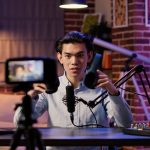 Asian content creator recording a podcast and reviewing headphones on camera in a home studio setup with microphone and laptop