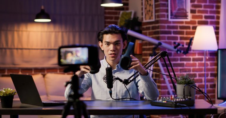 Asian content creator recording a podcast and reviewing headphones on camera in a home studio setup with microphone and laptop
