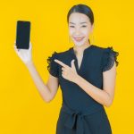 Smiling Asian woman holding and pointing at smartphone against yellow background, promoting a mobile app or digital service