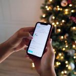 Person checking stock trading app with candlestick charts on smartphone beside a lit Christmas tree, mobile investing concept