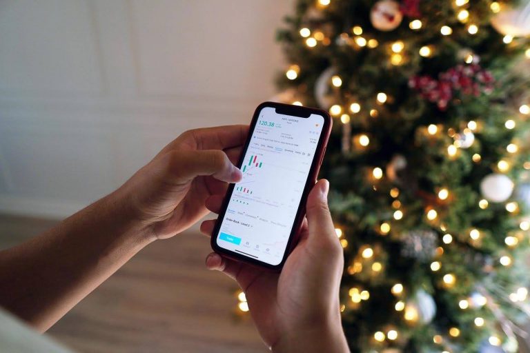 Person checking stock trading app with candlestick charts on smartphone beside a lit Christmas tree, mobile investing concept