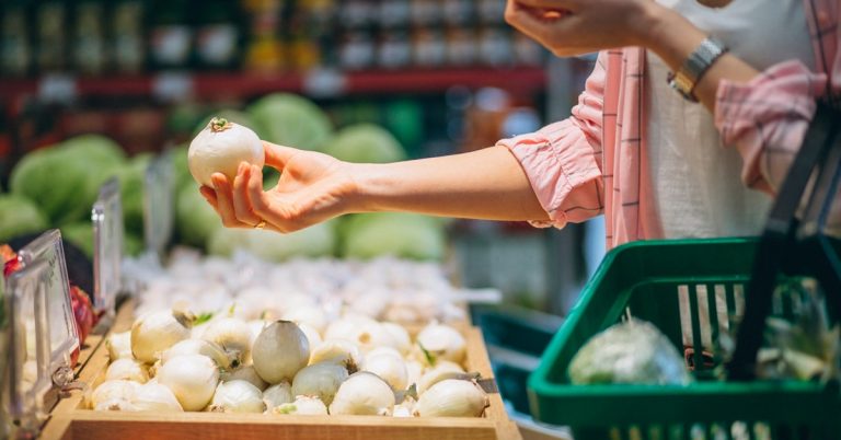 Woman selecting fresh onions at grocery store produce section while holding a shopping basket
