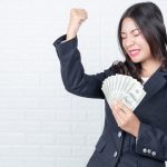 Businesswoman celebrating financial success while holding cash, raising fist in victory against white brick wall background
