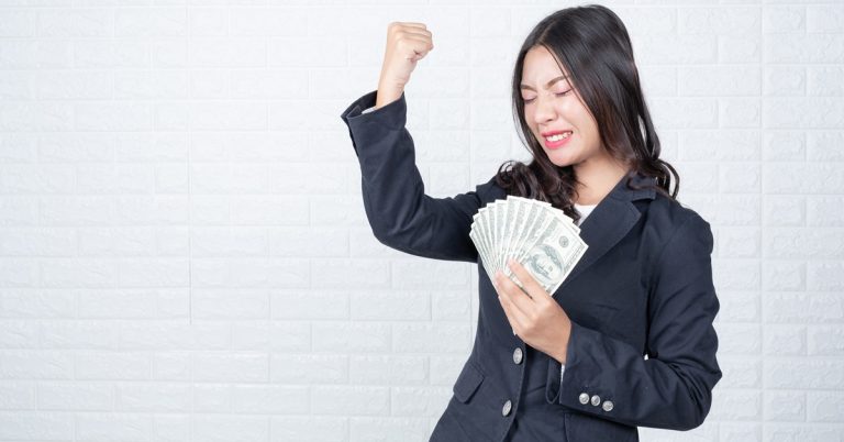 Businesswoman celebrating financial success while holding cash, raising fist in victory against white brick wall background