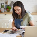 Woman calculating household expenses with calculator and bills at kitchen table, managing personal budget and finances