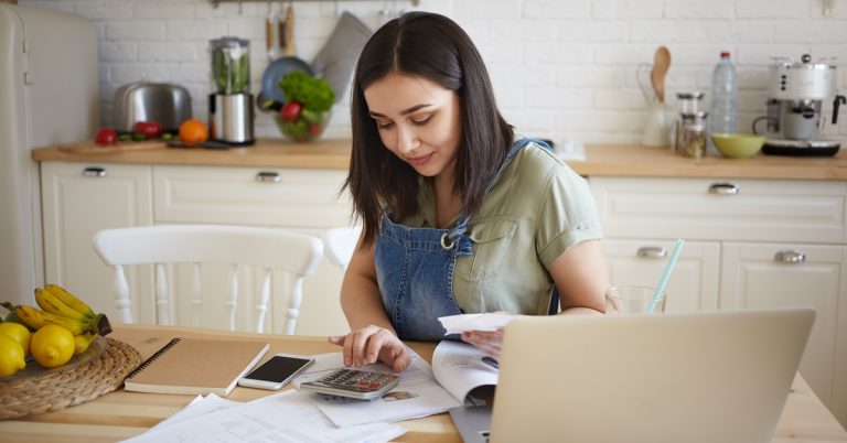 Woman calculating household expenses with calculator and bills at kitchen table, managing personal budget and finances