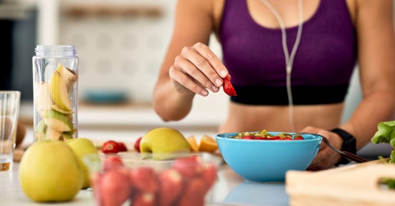 Athletic woman preparing healthy fruit salad with strawberries and apples in kitchen, promoting clean eating and fitness lifestyle