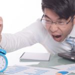 Frustrated office worker shouting at alarm clock while working late on laptop with documents spread across desk