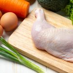 Raw chicken leg on cutting board with eggs, carrot, broccoli and fresh vegetables on white wooden surface