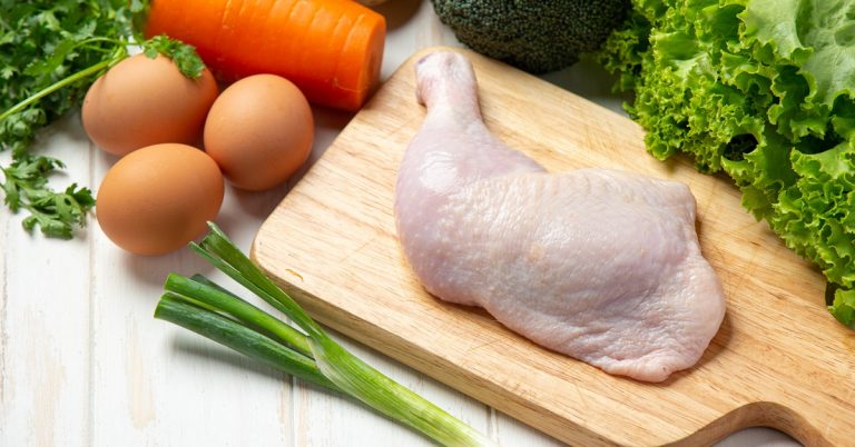 Raw chicken leg on cutting board with eggs, carrot, broccoli and fresh vegetables on white wooden surface
