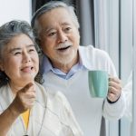 Happy Asian senior couple smiling and looking out window at home, enjoying retirement lifestyle and quality time together