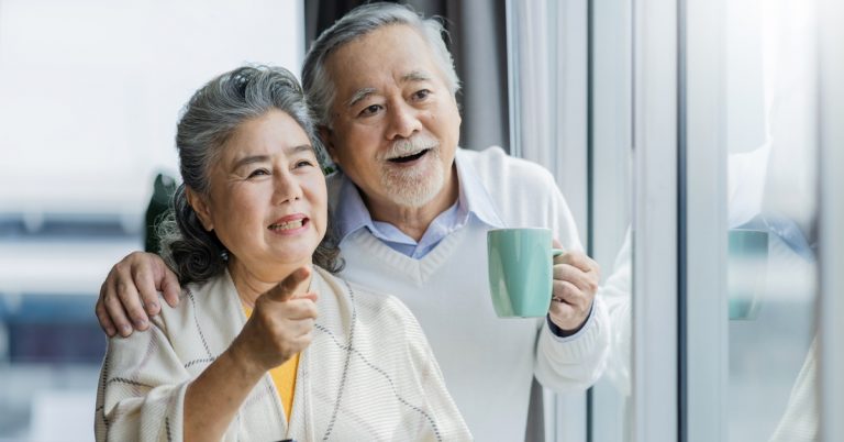 Happy Asian senior couple smiling and looking out window at home, enjoying retirement lifestyle and quality time together