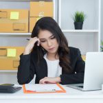 Small business owner reviewing paperwork at desk with laptop and packages, managing ecommerce orders and finances at home office