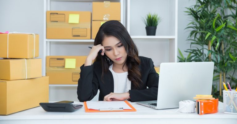 Small business owner reviewing paperwork at desk with laptop and packages, managing ecommerce orders and finances at home office
