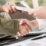 Business handshake sealing deal with document exchange over laptop and financial charts, symbolizing agreement and partnership