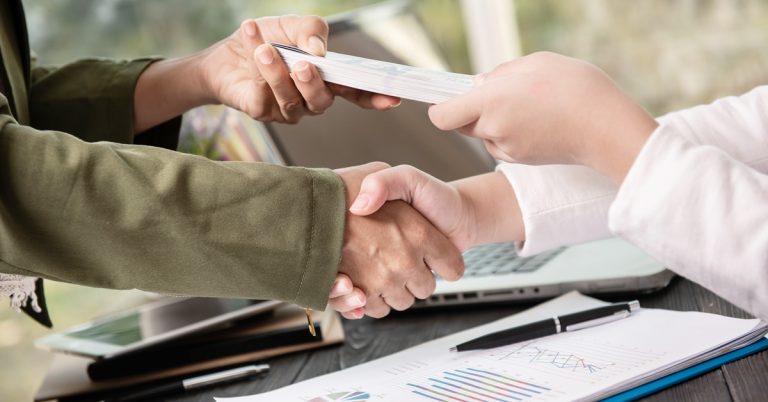 Business handshake sealing deal with document exchange over laptop and financial charts, symbolizing agreement and partnership