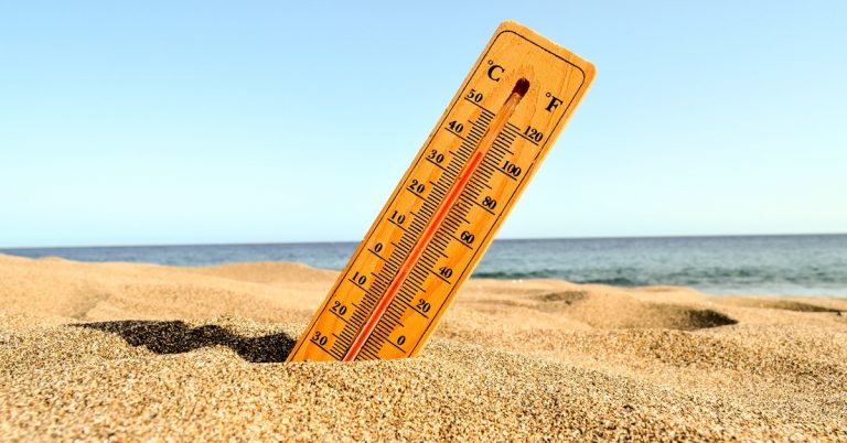 Thermometer in beach sand showing high temperature near ocean, symbolizing heatwave, summer weather and climate change