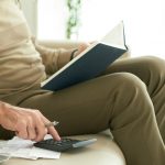 Person calculating expenses with calculator and notebook at home, reviewing bills, budgeting and managing personal finances