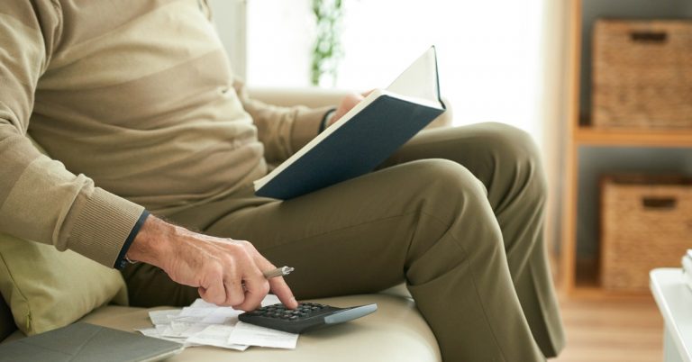 Person calculating expenses with calculator and notebook at home, reviewing bills, budgeting and managing personal finances