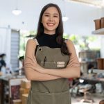 Smiling female ceramic artist standing in pottery workshop with handmade clay products, representing small business and craftsmanship