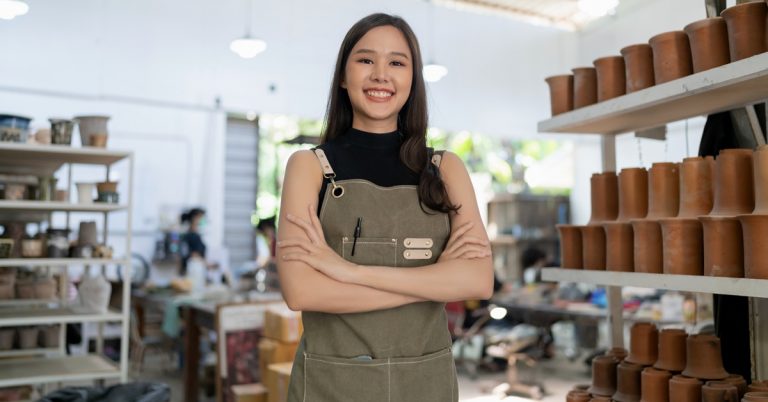 Smiling female ceramic artist standing in pottery workshop with handmade clay products, representing small business and craftsmanship