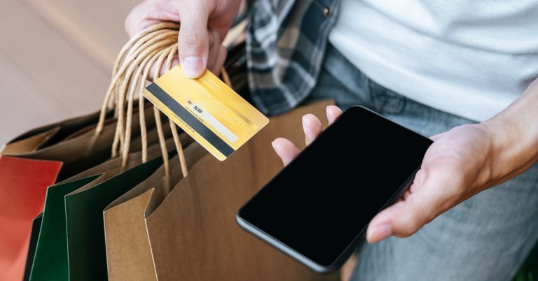 Person shopping with credit card and smartphone while carrying retail bags, representing cashless consumer spending